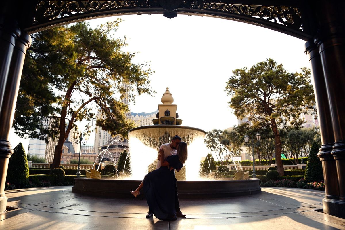 A couple sharing a dip kiss in front of a garden fountain, framed by an ornate dark archway in late afternoon sun