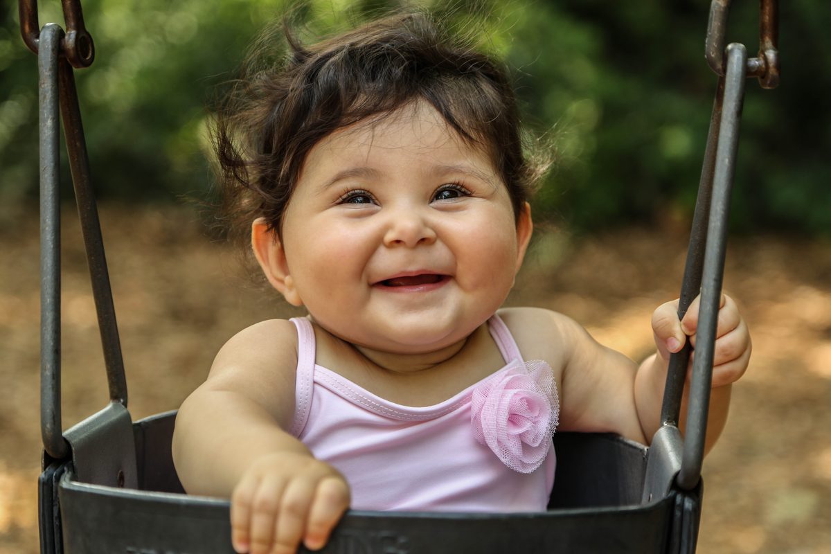 A laughing baby girl in a pink top holding on to a park swing