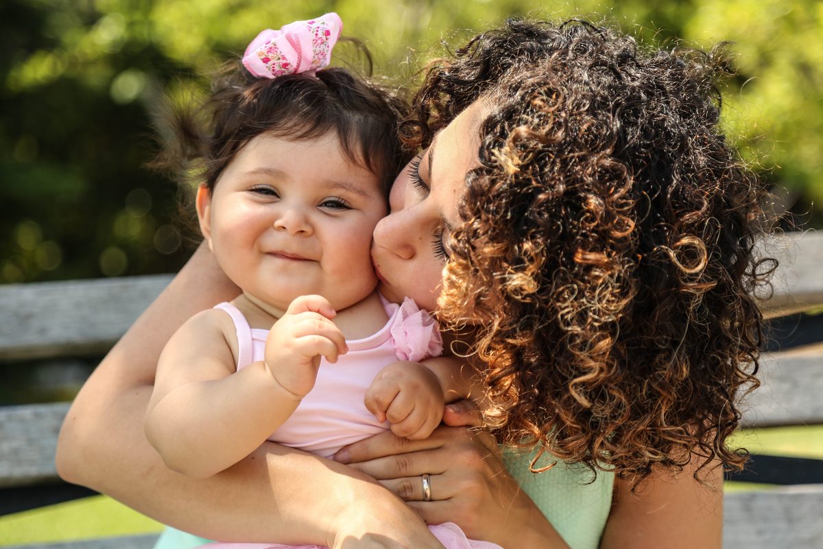 A mom with curly hair kissing her smiling baby girl on the cheek, outdoors in summer sun